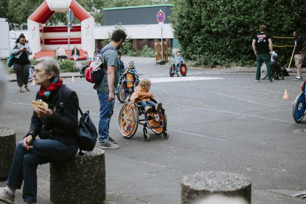 Kinder probieren Rollstühle aus und spielen Rollstuhlbasketball im Hintergrund ist die Hüpfburg zu sehen